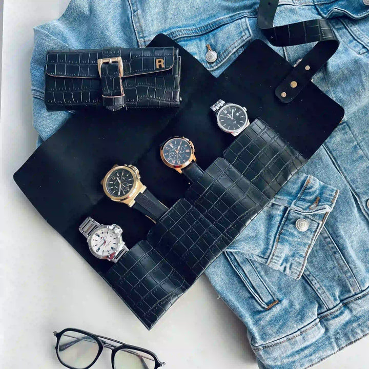 A black crocodile-embossed watch roll organizer laid flat on a light surface, holding several wristwatches, with a matching black watch pouch and a light blue denim jacket nearby. No person or real environment is present, so this is Product/Packaging rather than Lifestyle.