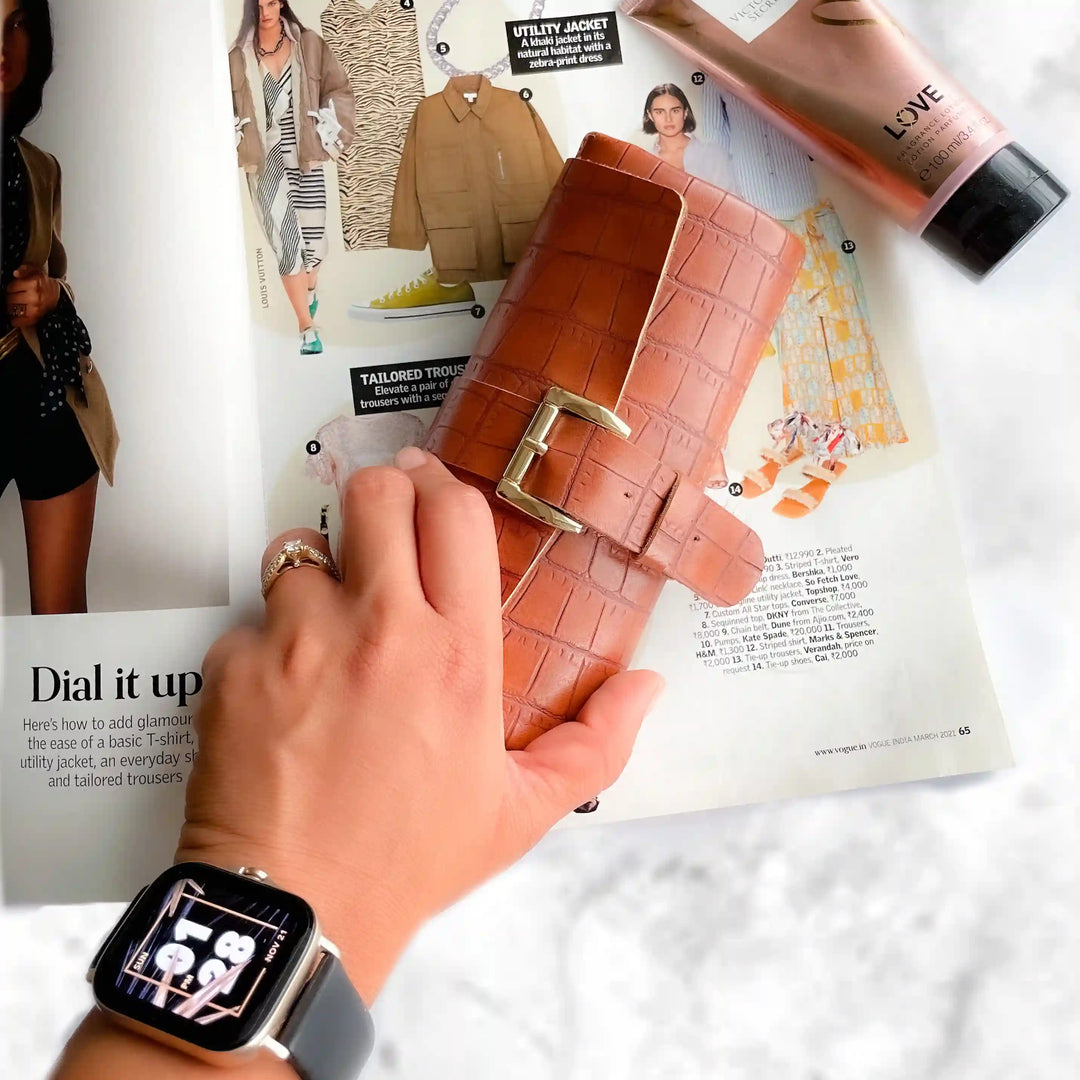 A brown crocodile-embossed leather wallet with a gold buckle, held by a hand over an open magazine page with fashion items and a pink tube of lip gloss.