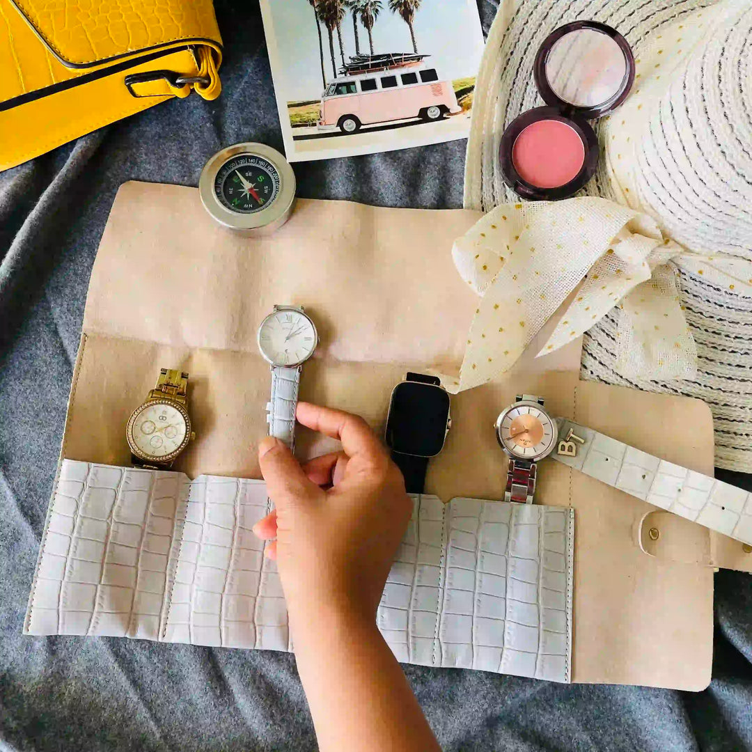 A grey travel watch roll-up organizer bag laid flat on a gray fabric surface, holding several watches. A hand is placing a watch into the center slot.