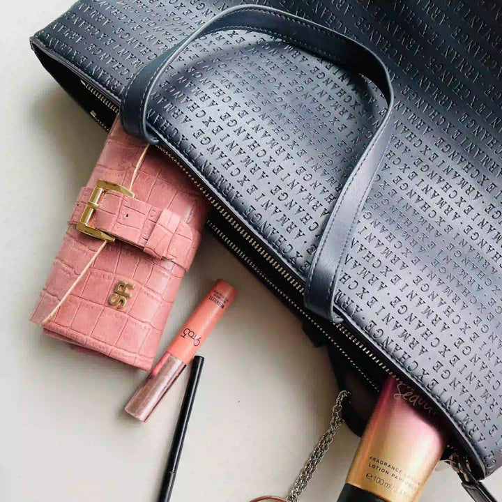 A black textured zippered handbag with matching shoulder strap, shown alongside a pink crocodile-embossed wallet, a pink lip product, and other cosmetics on a light surface.