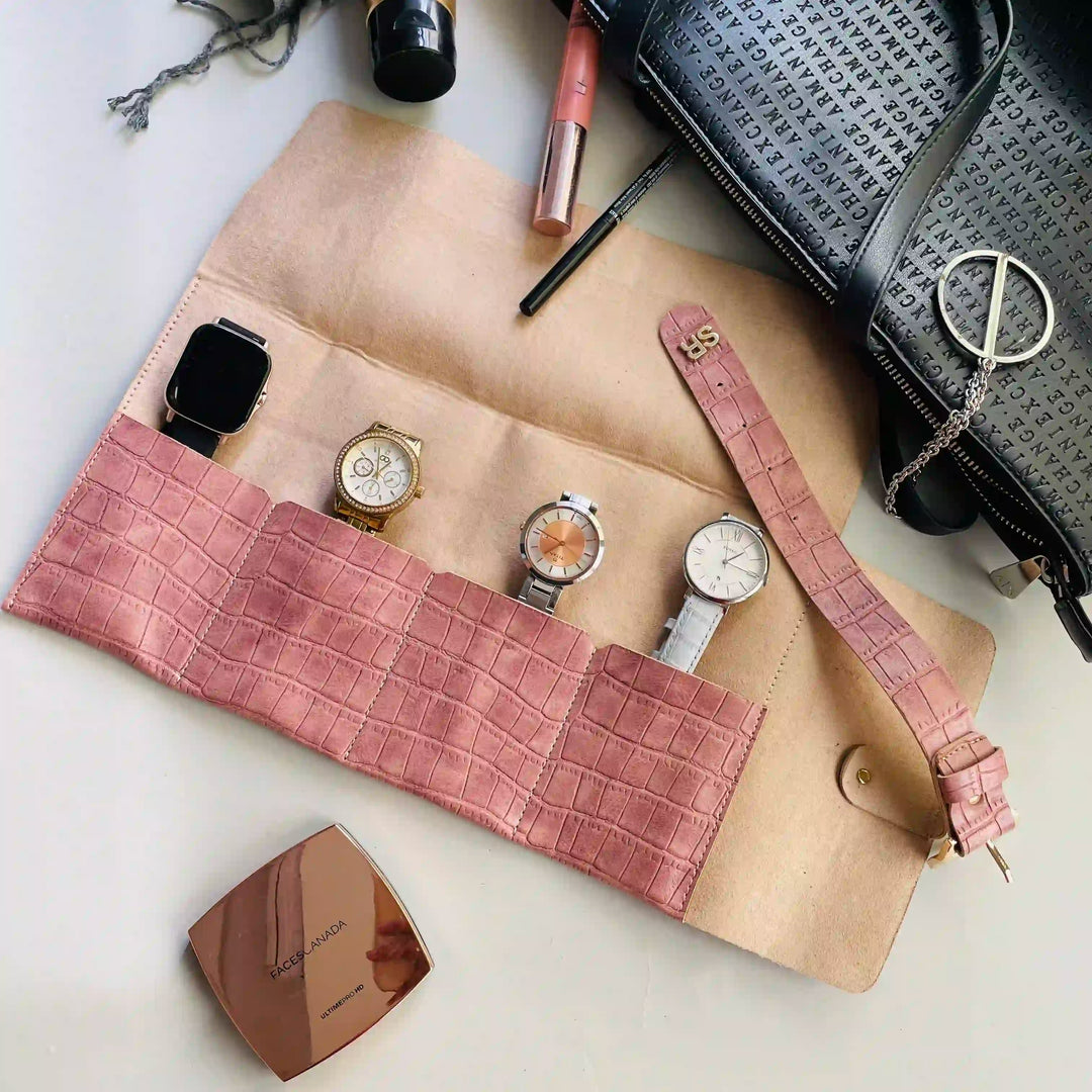 A onion pink crocodile-embossed watch roll organizer laid flat on a light surface, holding four watches. The scene is styled with a black patterned handbag, a rose gold compact mirror, and pink and black cosmetics.