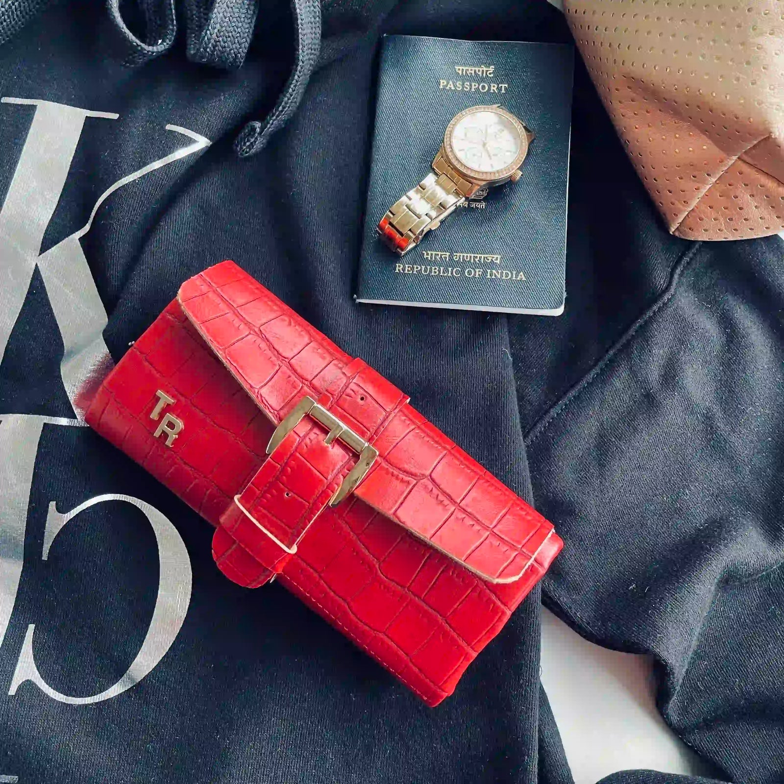 A bright red crocodile-embossed leather wallet with a gold-tone buckle, styled on a black hoodie next to a passport and a watch.