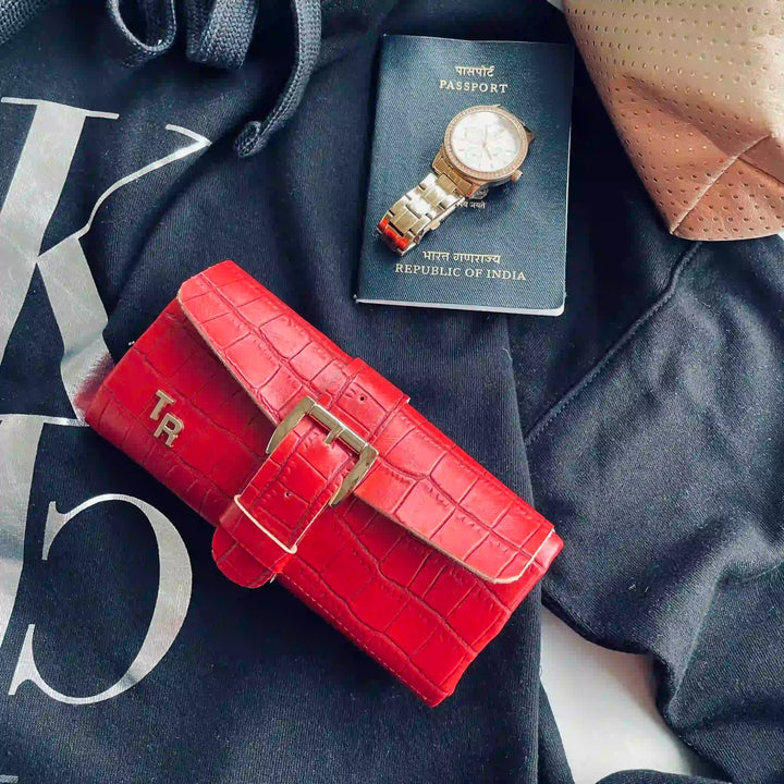 A bright red crocodile-embossed leather wallet with a gold-tone buckle, styled on a black hoodie next to a passport and a watch.