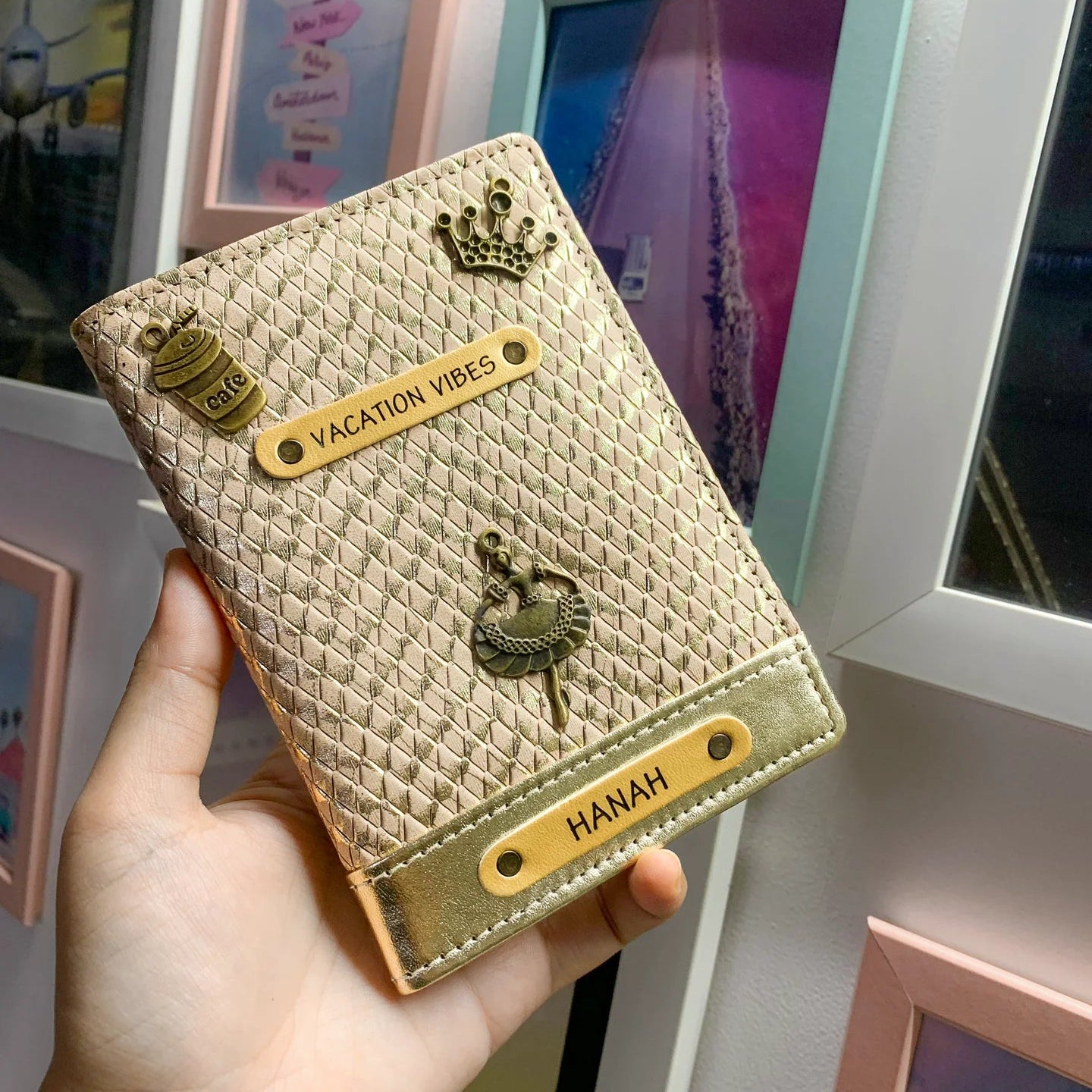 A personalized passport cover in rose gold color, featuring a name tag and charm, displayed in a hand.