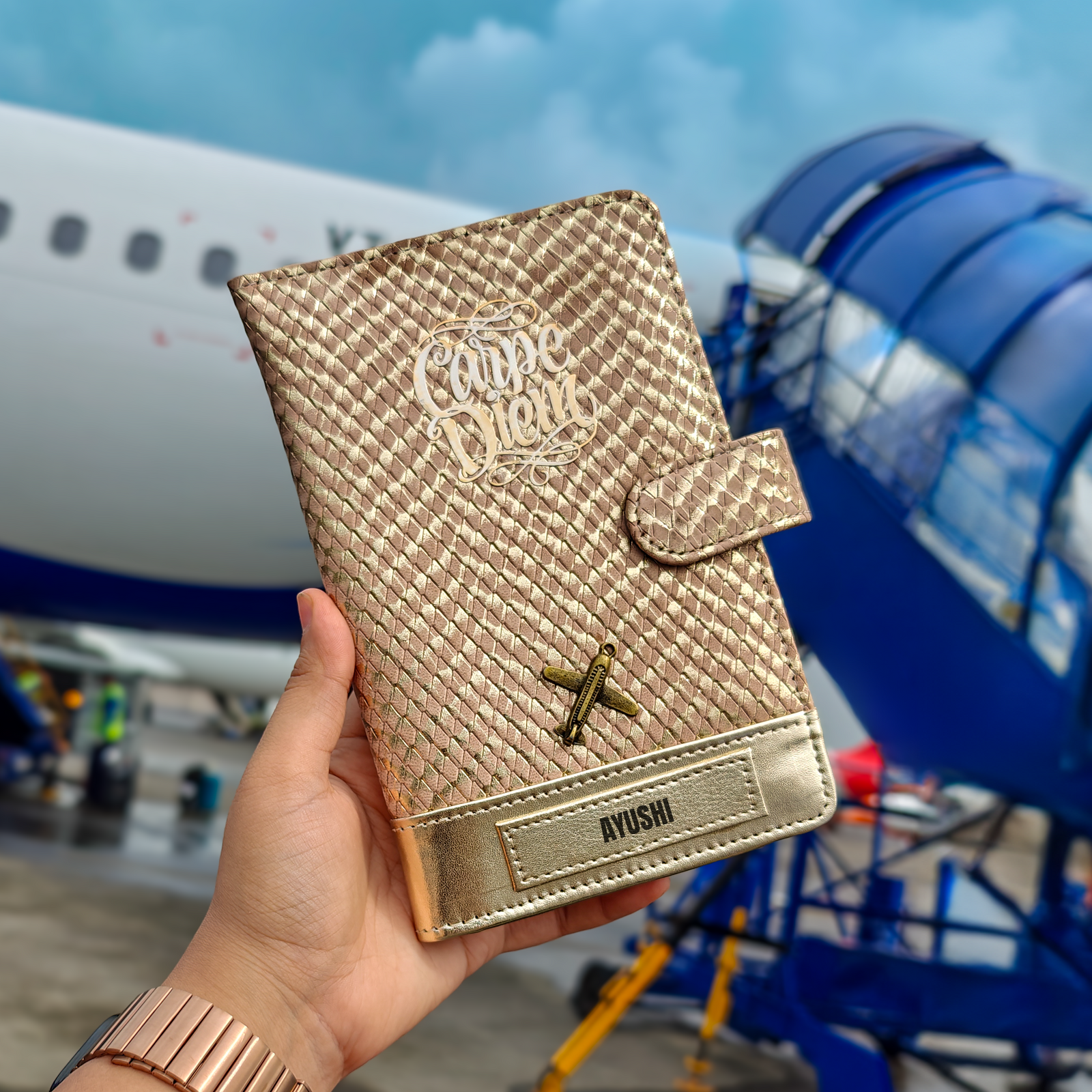 A person holding a rose gold travel wallet by TPC Gifts with a designer exterior, showing a personalized name tag and an airplane window in the background.