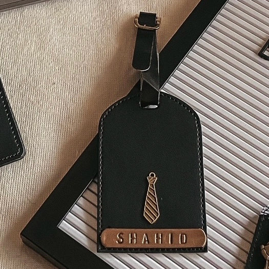 A black leather luggage tag with a gold-tone tie emblem and a custom nameplate reading &