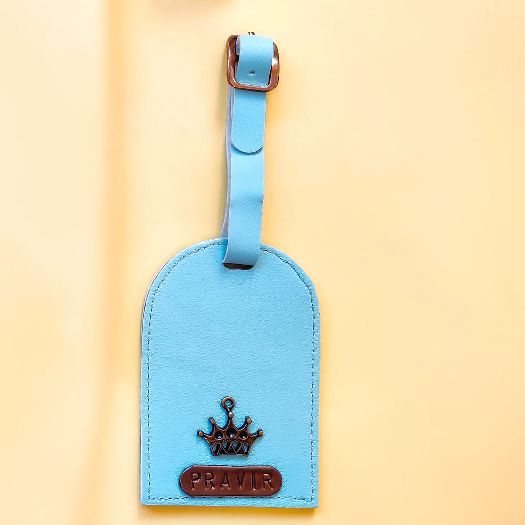 A light blue leather luggage tag with a matching strap and a brown crown emblem, featuring the name &