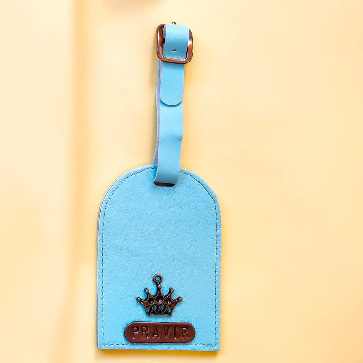A light blue leather luggage tag with a matching strap and a brown crown emblem, featuring the name &