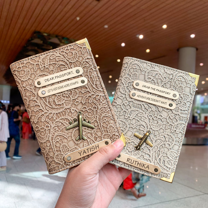 Two people holding black and white passport covers by TPC Gifts with custom text and charms, displayed in an airport setting.