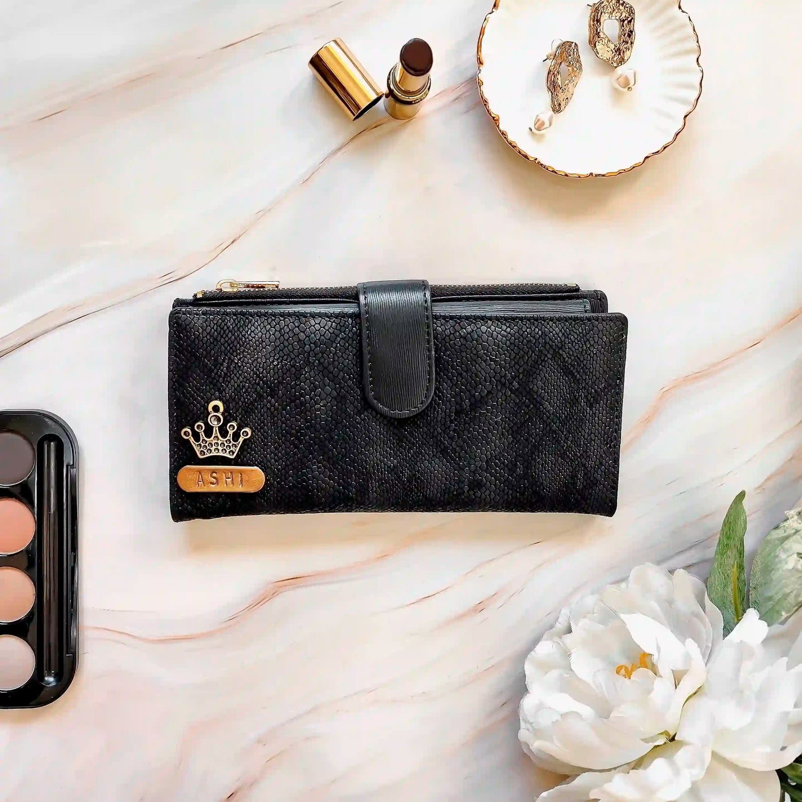Black wallet with a crown emblem and 'ASH' on a marble surface with flowers and accessories.