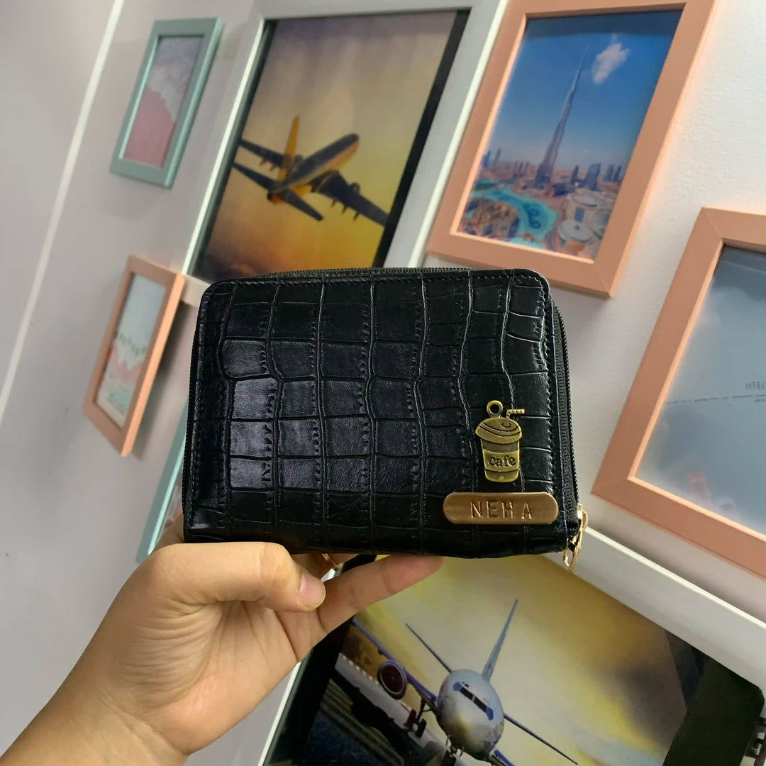 Black crocodile-patterned wallet held in front of framed pictures on a wall.