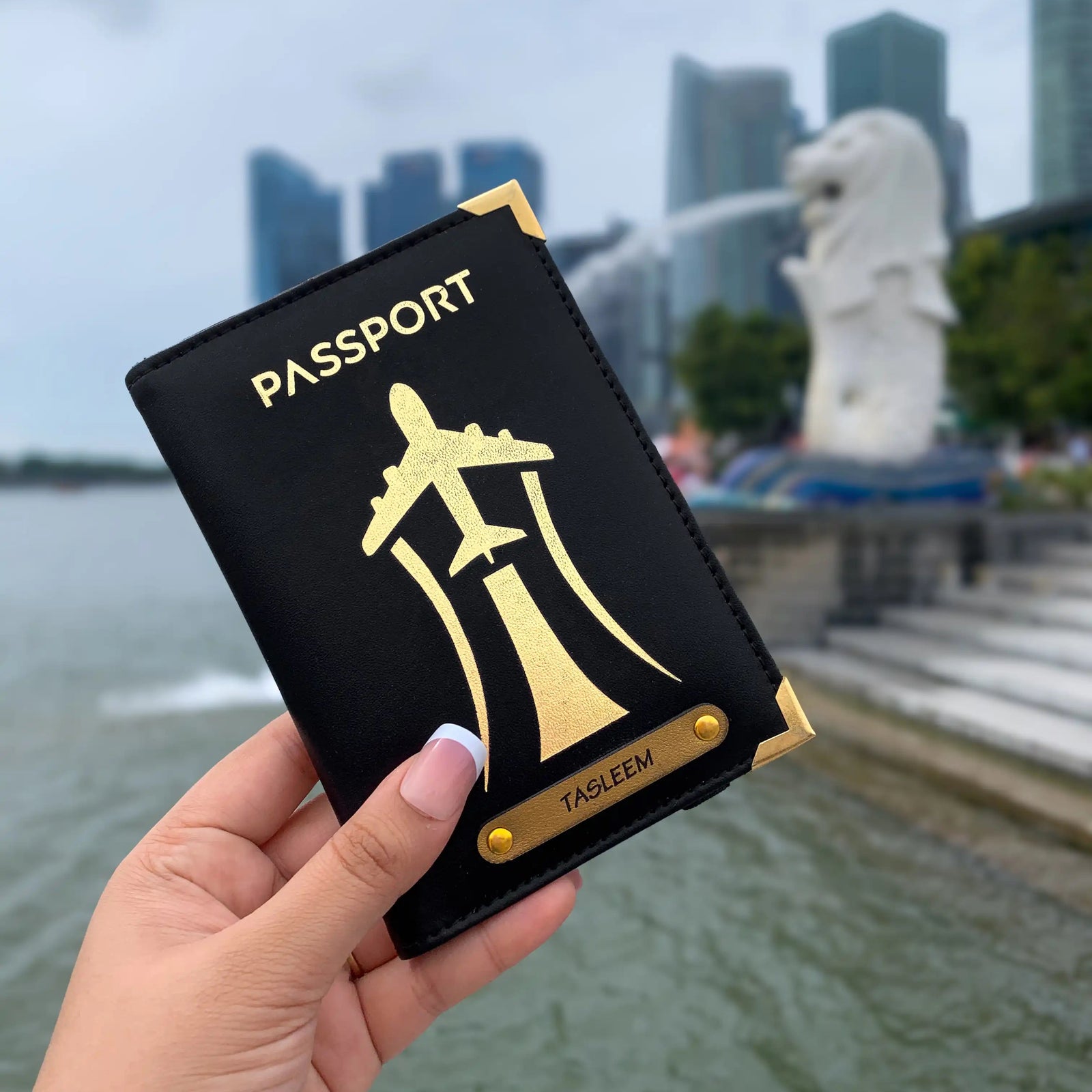 A hand holding a black passport cover with gold aviation-inspired design and text 'PASSPORT'. The cover is displayed in front of a blurred background featuring a statue and buildings.