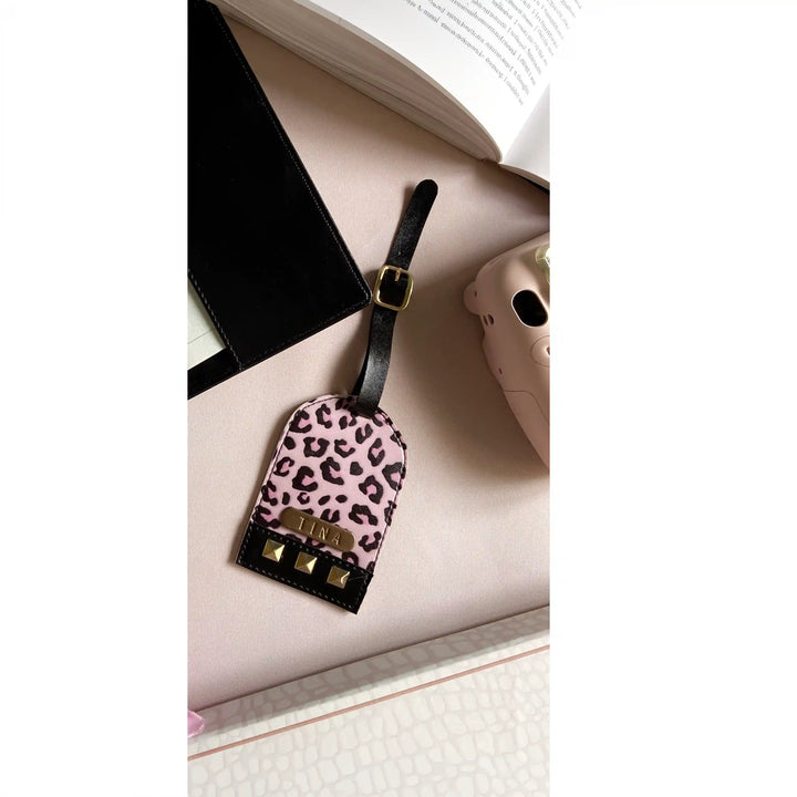A pink leopard-print luggage tag with black leather trim and gold hardware, styled on a light pink surface next to an open book and a beige phone case.