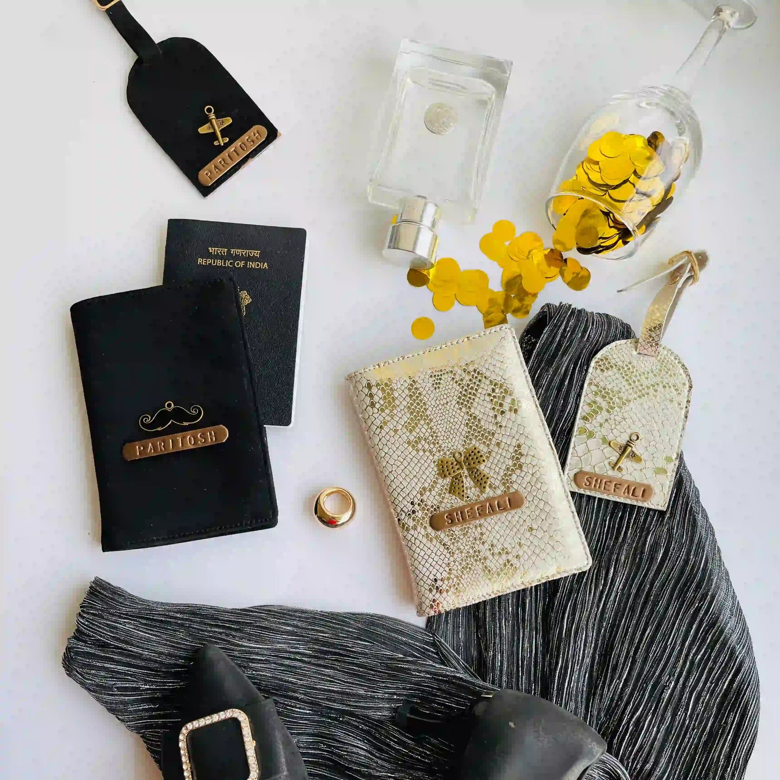 A styled flat-lay of travel accessories on a white surface, including two black passport covers with gold lettering, a black luggage tag, a cream snakeskin-patterned wallet or card holder with a gold logo, a cream luggage tag, a clear perfume bottle, a glass bottle with gold confetti, scattered gold coins, and a black-and-gray pleated fabric runner with black shoes partially visible.