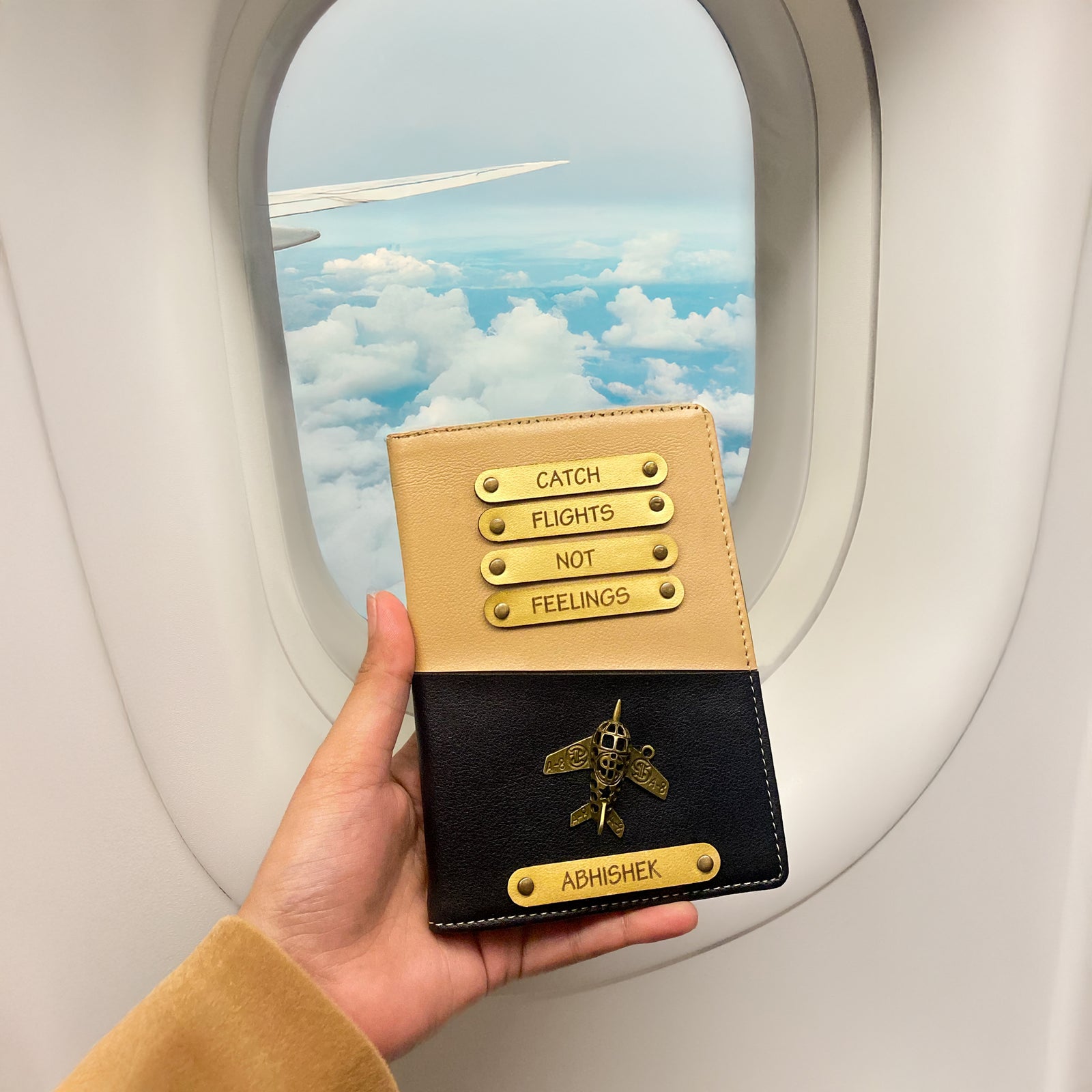 A person holding a dual tone black and beige passport cover with personalized text and a logo, while on an airplane with a cloudy sky outside the window.