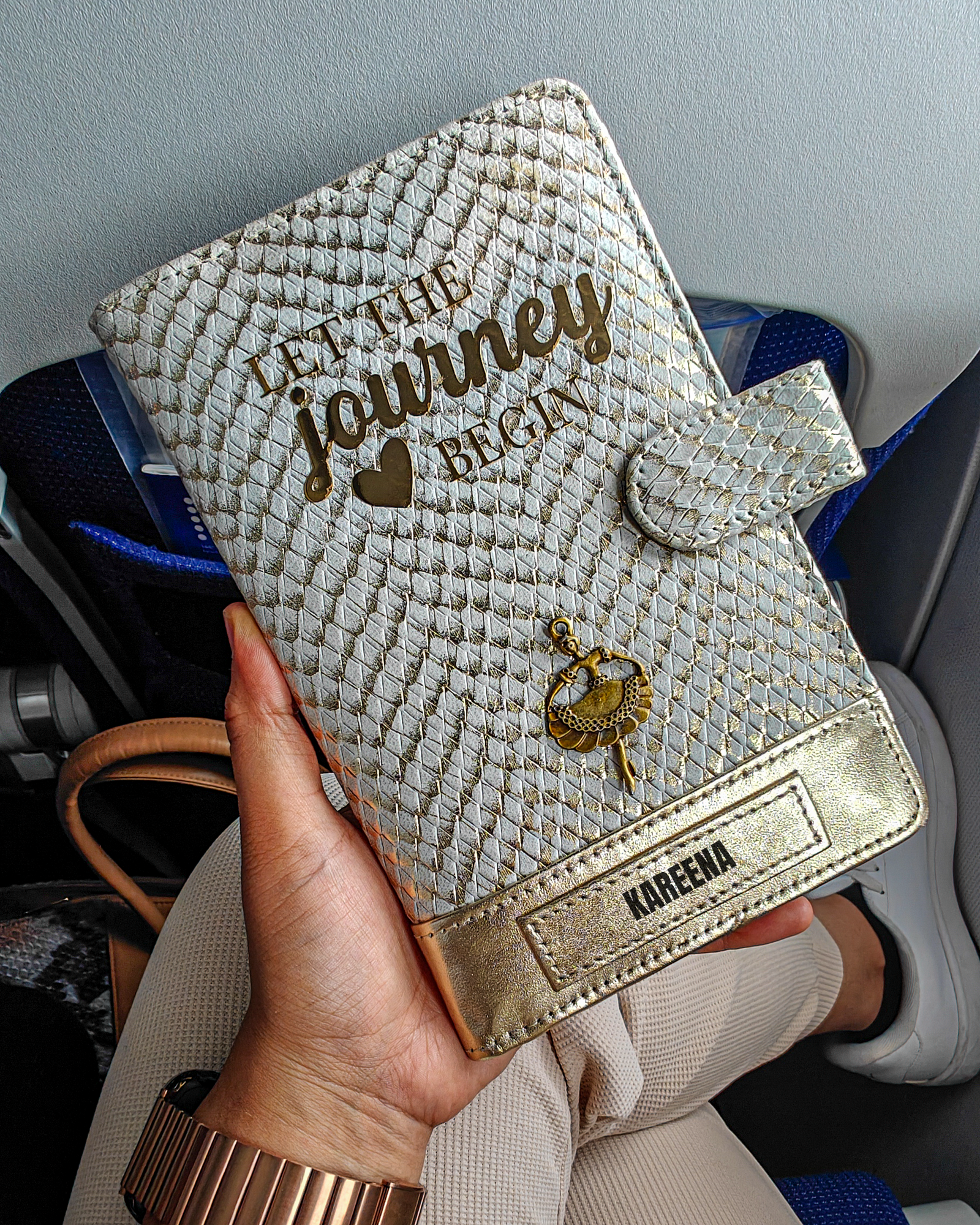 A personalized travel wallet by TPC Gifts with gold and ivory colors, featuring text and a crest design, held in a person's hand while on an airplane.