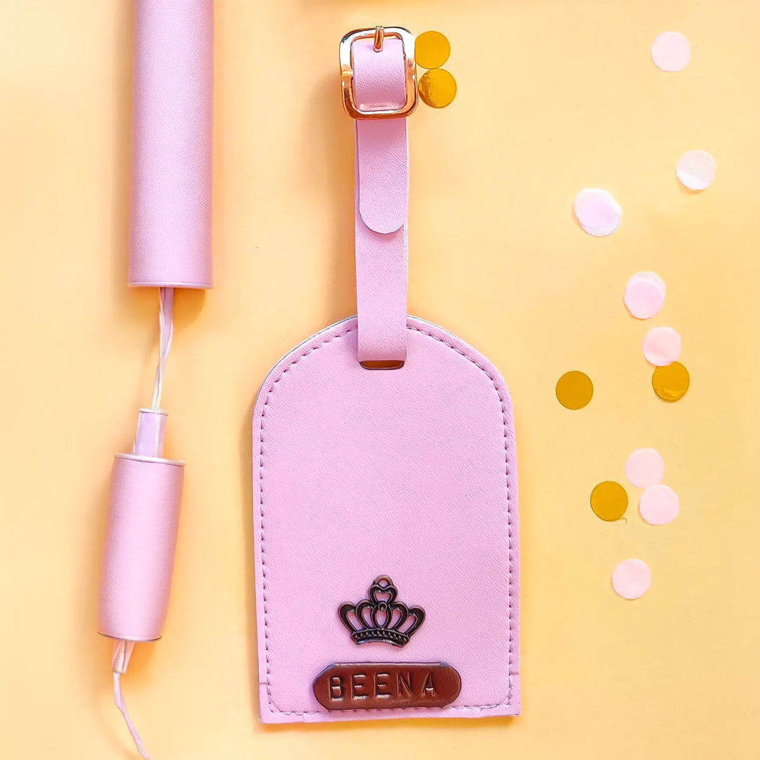 A light pink leather luggage tag with a gold-tone buckle and a metal crown emblem featuring the text &