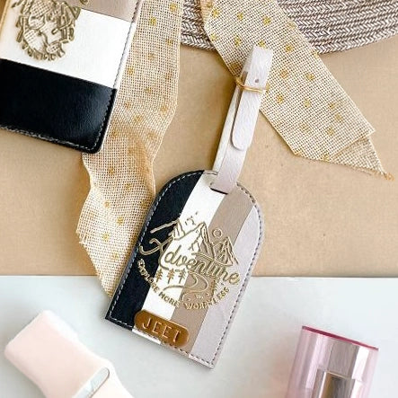 A leather luggage tag with a diagonal black, beige, and white stripe design, featuring gold foil embossed text that reads &