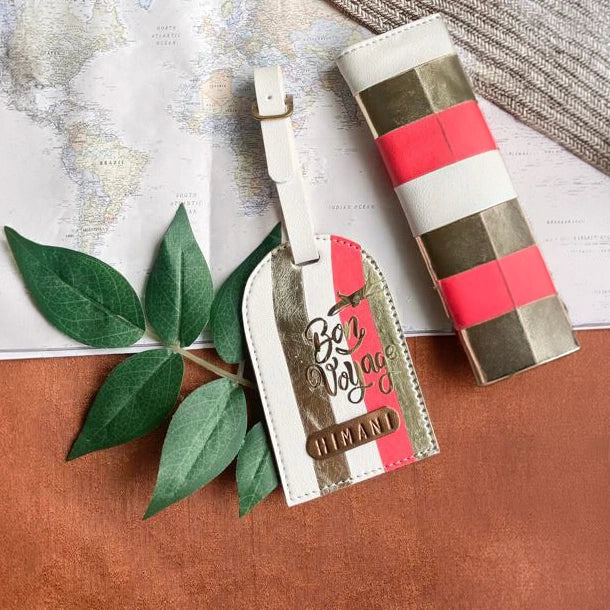 A luggage tag and matching cylindrical pouch with a striped pattern of metallic gold, white, and coral pink, styled on a map background with green leaves and a brown textured surface.