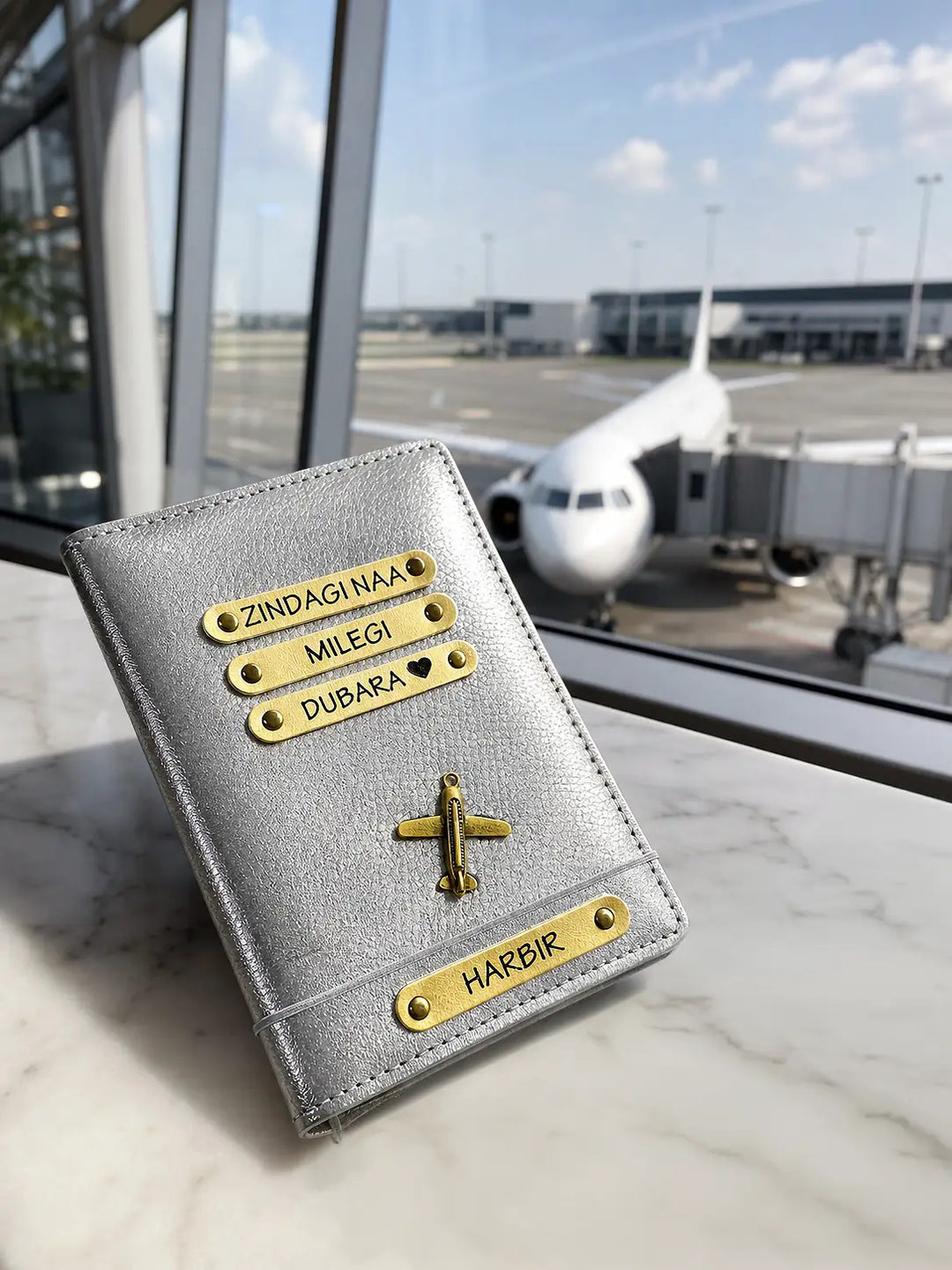 Silver passport holder with gold text and accents on a surface with an airplane in the background from tpc gifts