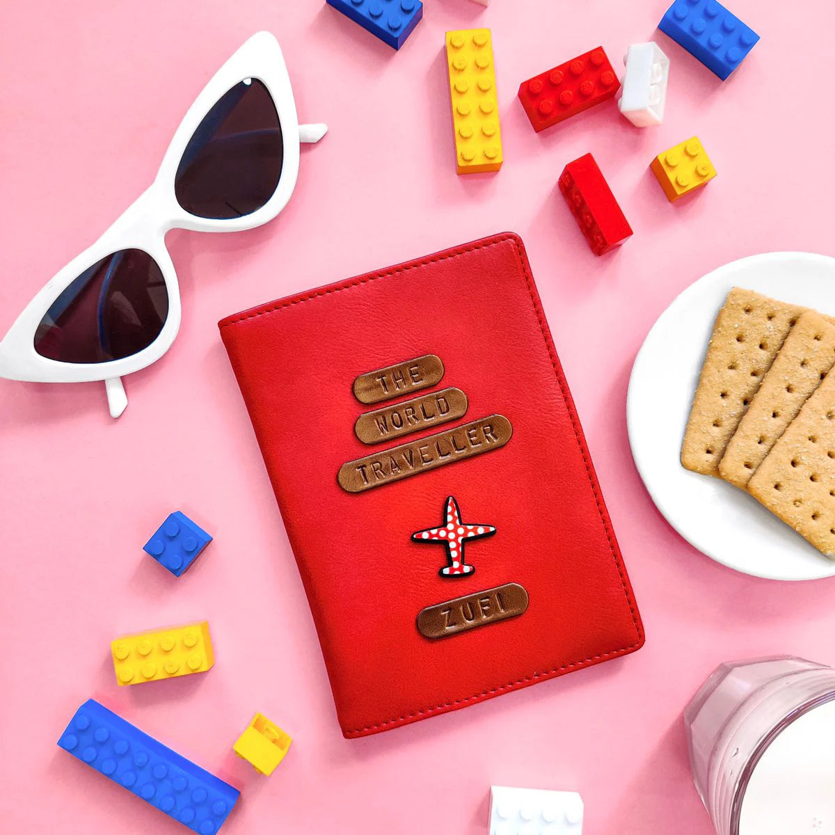Personalised Red passport holder with  a name and charm & quote 'The World Traveler' design on a pink background with toys and snacks with playful background setup  - TPC Gifts