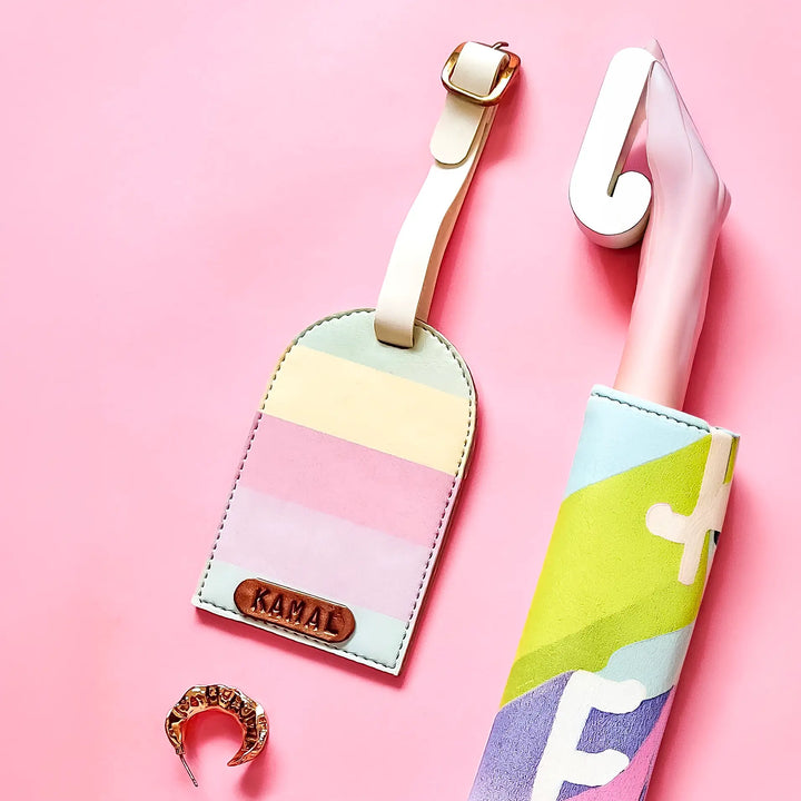 A pastel striped luggage tag with a cream strap and a gold-toned KANAL logo plate, shown next to a pink and white umbrella in a matching geometric print, on a solid pink background.