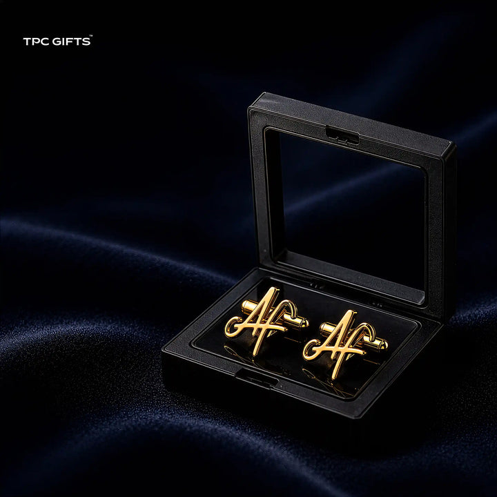 A pair of gold cufflinks shaped like intertwined script letters, presented in an open black gift box with a clear lid, set against a dark navy satin fabric background. The brand name &