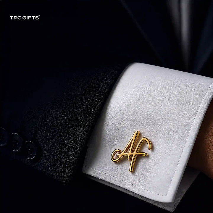 A close-up of a white dress shirt cuff pinned with a gold script monogram pin reading &