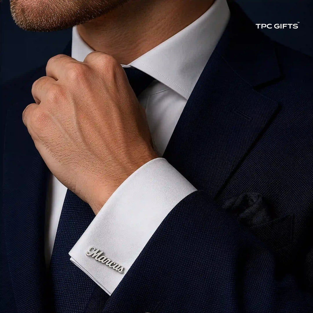 A close-up of a man wearing a navy suit, white dress shirt, and dark tie, with his hand adjusting the collar. A silver script cufflink is visible on the cuff.