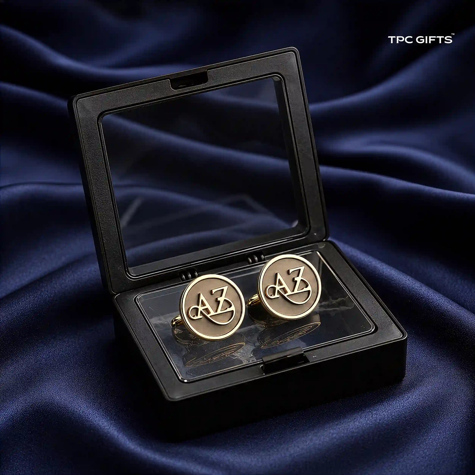 A pair of round gold cufflinks with interlocking 'AZ' lettering, presented in an open black gift box with a clear lid, set against a dark blue satin fabric background. The 'TPC GIFTS' logo is visible in the top right corner.