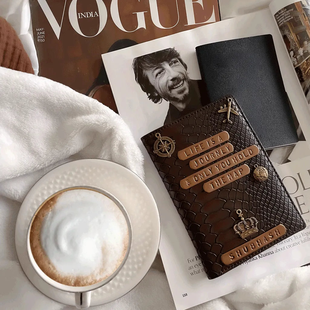 Personalised Passport Cover Men - Dark Brown Croco Texture with embossed text and gold-tone charms, styled on top of open Vogue India magazine next to a cup of latte macchiato on a white blanket - TPC Gifts