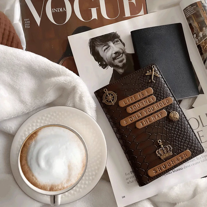 Personalised Passport Cover Men - Dark Brown Croco Texture with embossed text and gold-tone charms, styled on top of open Vogue India magazine next to a cup of latte macchiato on a white blanket - TPC Gifts