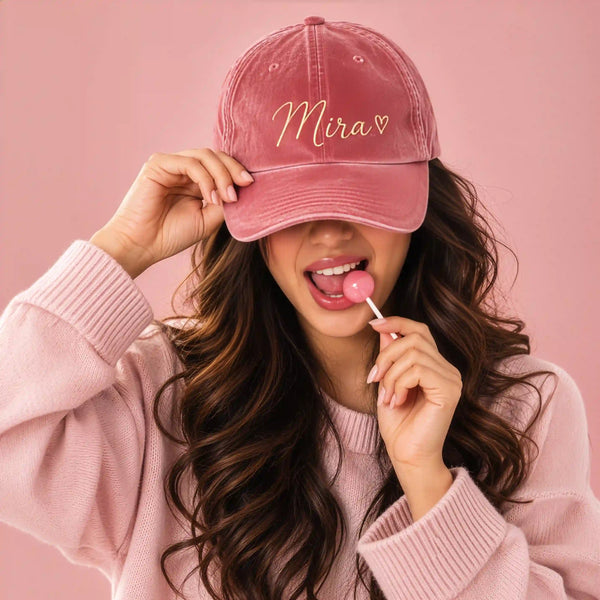 Woman wearing a pink velvet baseball cap with the name "Mira" and a heart embroidered in gold cursive thread
