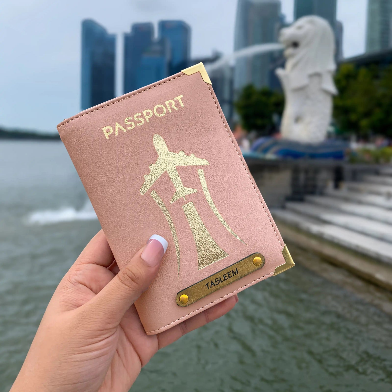 A hand holding a black passport cover with gold aviation-inspired design and text 'PASSPORT'. The cover is displayed in front of a blurred background featuring a statue and buildings.