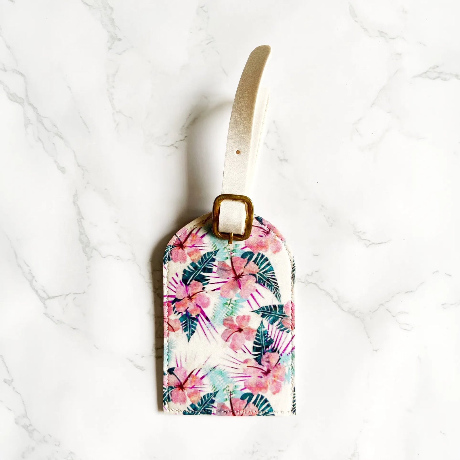 A rectangular luggage tag with a tropical floral and palm leaf print, featuring a cream-colored strap and a gold metal buckle, displayed on a white marble surface.