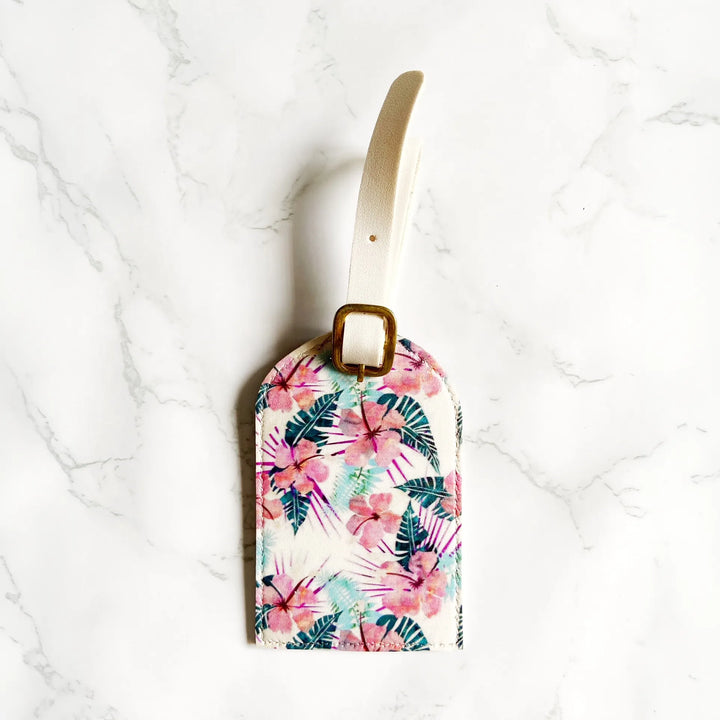 A rectangular luggage tag with a tropical floral and palm leaf print, featuring a cream-colored strap and a gold metal buckle, displayed on a white marble surface.