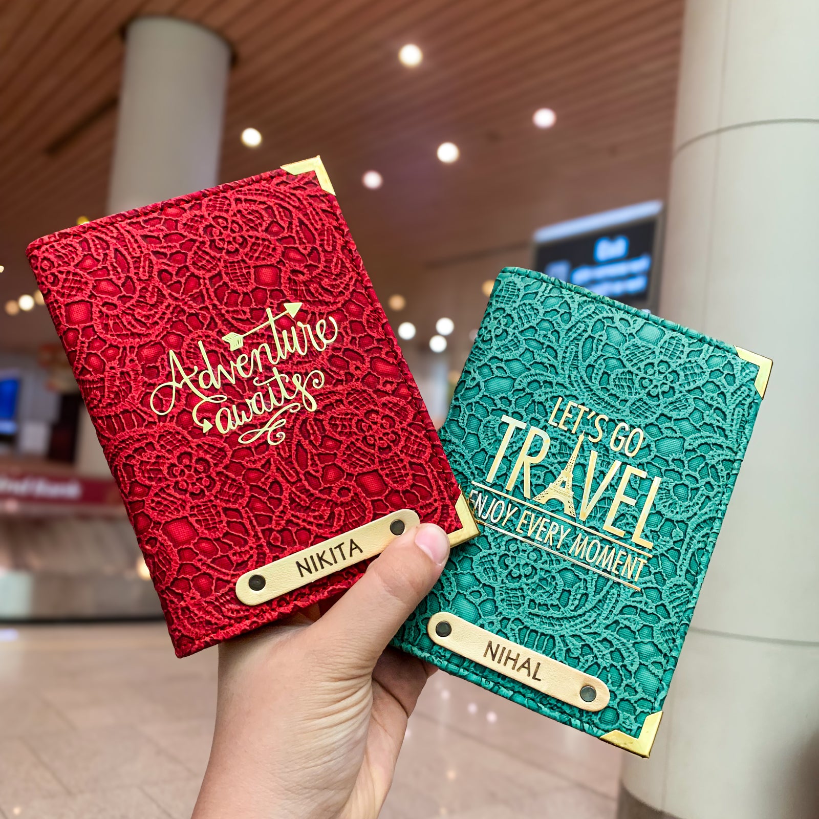 Two people holding black and white passport covers with custom text and charms, displayed in an airport setting.