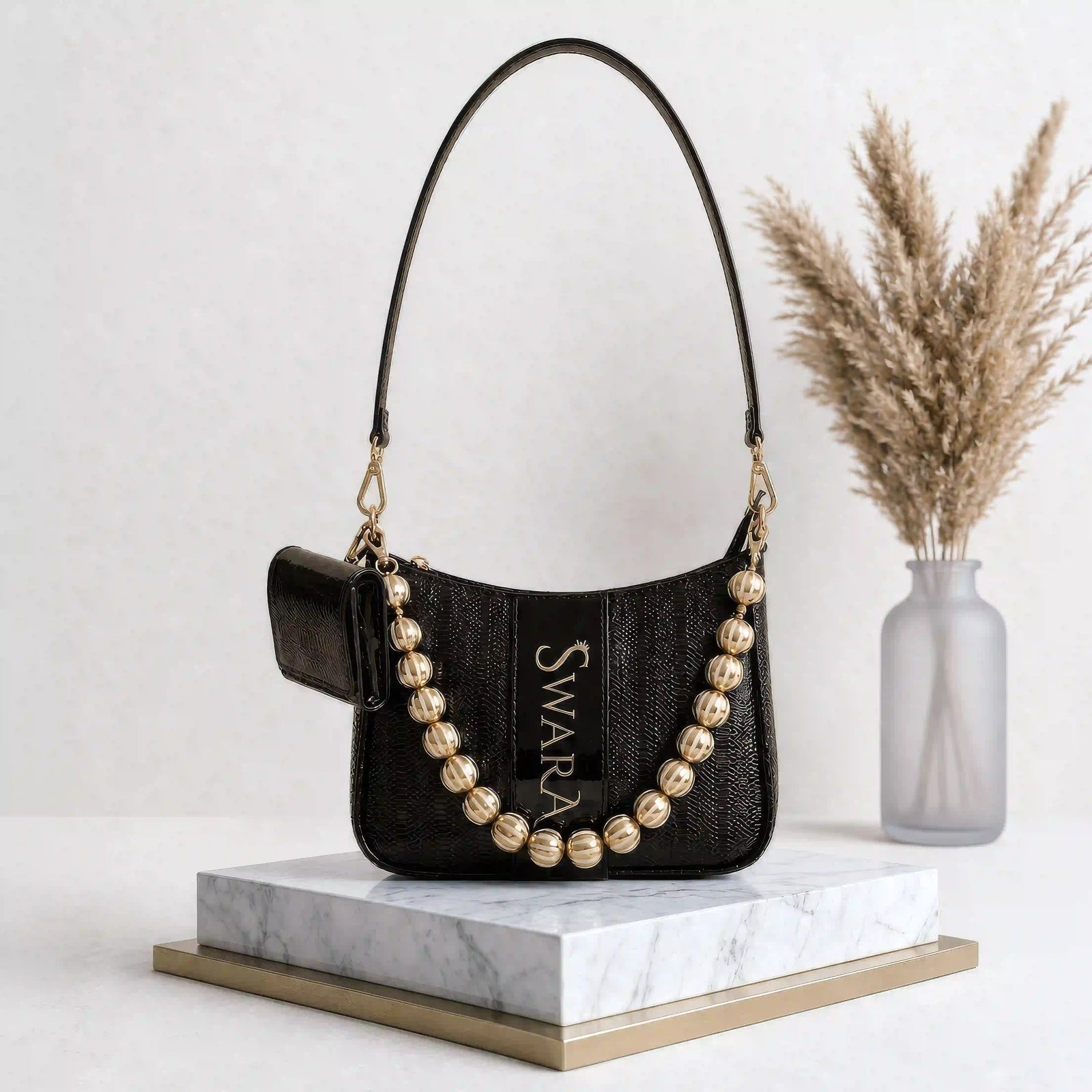 Black personalized sling bag by TPC Gifts  with gold beads on a marble stand with a vase of dried plants in the background.