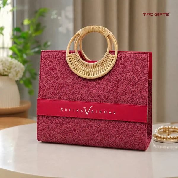 A structured magenta-red textured tote bag with a circular woven rattan handle, featuring a red horizontal band with the brand name 'RUPIKA VAIBHAV' in white. The bag is displayed on a light-colored table with a blurred indoor background.