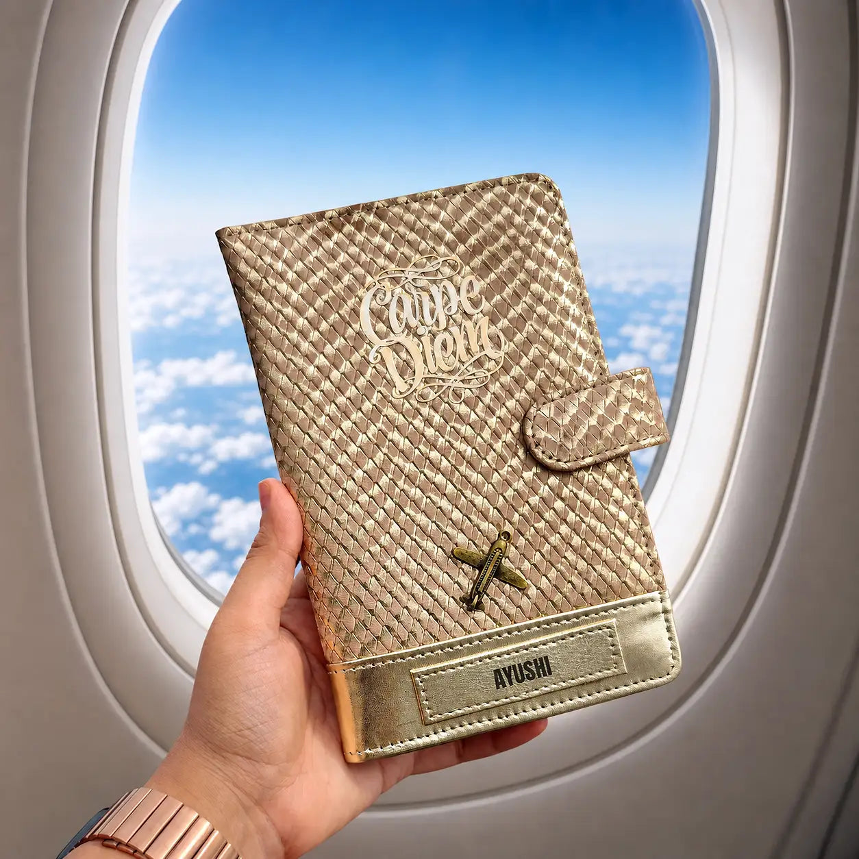 Hand holding a rose gold travel wallet with 'Carpe diem' in front of an airplane window with a blue sky view - TPC Gifts