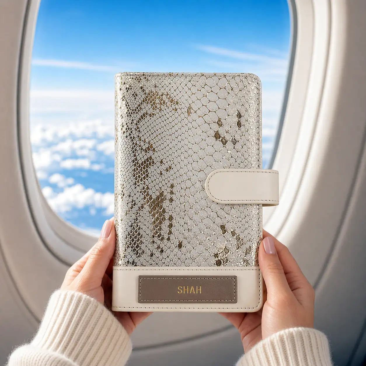 Hand holding a textured notebook with 'SHAH' name in front of an airplane window with a view of clouds.