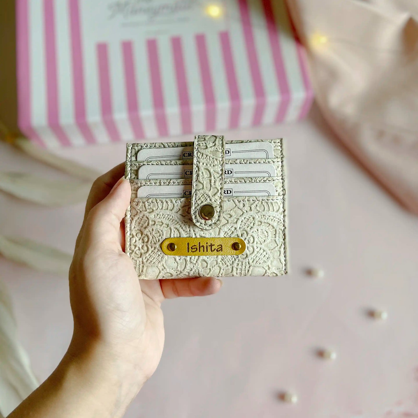 Hand holding a decorative wallet with 'Ishita'  against a pink and white striped background.