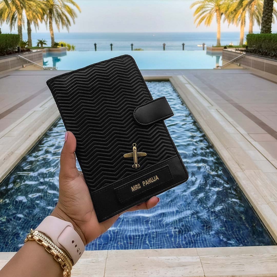 Black textured travel wallet or passport cover by TPC Gifts held by a hand with gold watch and bracelet, pool and palm trees in the background