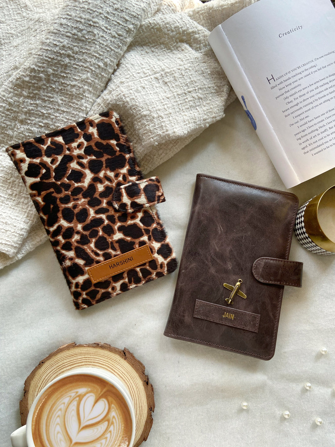 Two travel organizers, one with a solid brown color and another with a animal print, placed on a surface with a cup of coffee and a book at backgroud setup from TPC Gifts