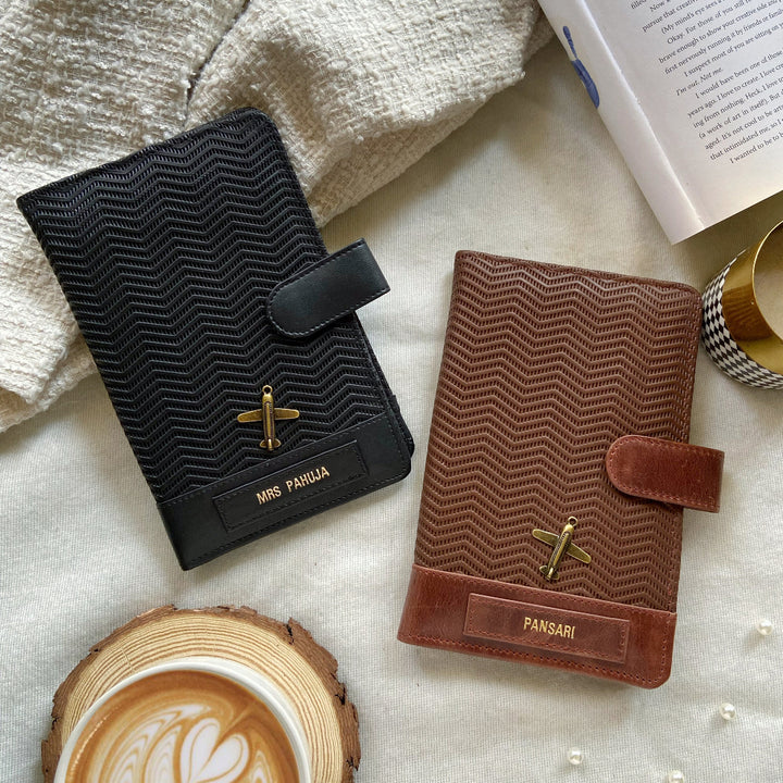 Two leather travel organizers laid flat, one black and one brown, with a wave pattern design, surrounded by a cup of coffee and a book.