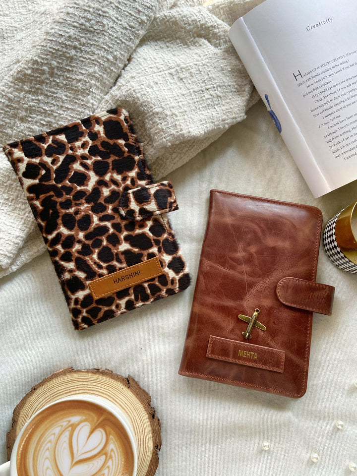 Two travel organizers, one with a solid brown color and another with a animal print, placed on a surface with a cup of coffee and a book at backgroud setup from TPC Gifts