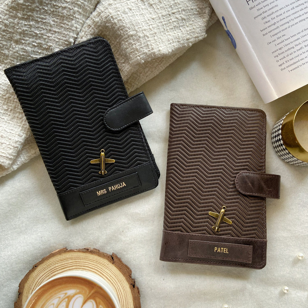 Two leather travel organizers laid flat, one black and one brown, with a wave pattern design, surrounded by a cup of coffee and a book.