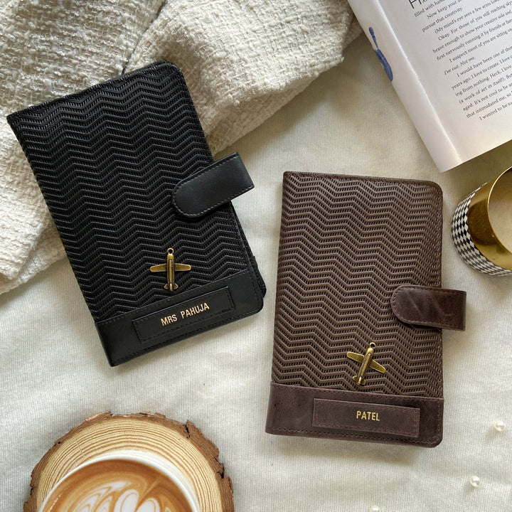 Two leather travel organizers laid flat, one black and one brown, with a wave pattern design, surrounded by a cup of coffee and a book.