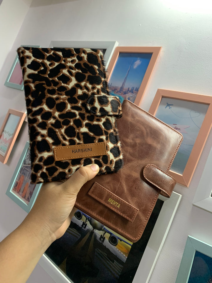 Two travel organizers, one with a solid brown color and another with a animal print, placed on a surface with a cup of coffee and a book at backgroud setup from TPC Gifts
