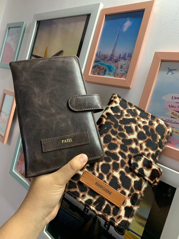 Two travel organizers, one with a solid brown color and another with a animal print, placed on a surface with a cup of coffee and a book at backgroud setup from TPC Gifts